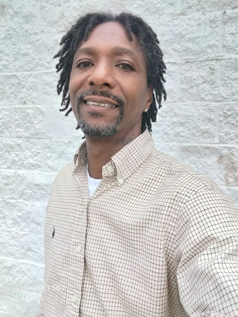 Portrait of the founder of EHI Handyman Services standing in front of a white brick wall, wearing a checkered dress shirt and smiling