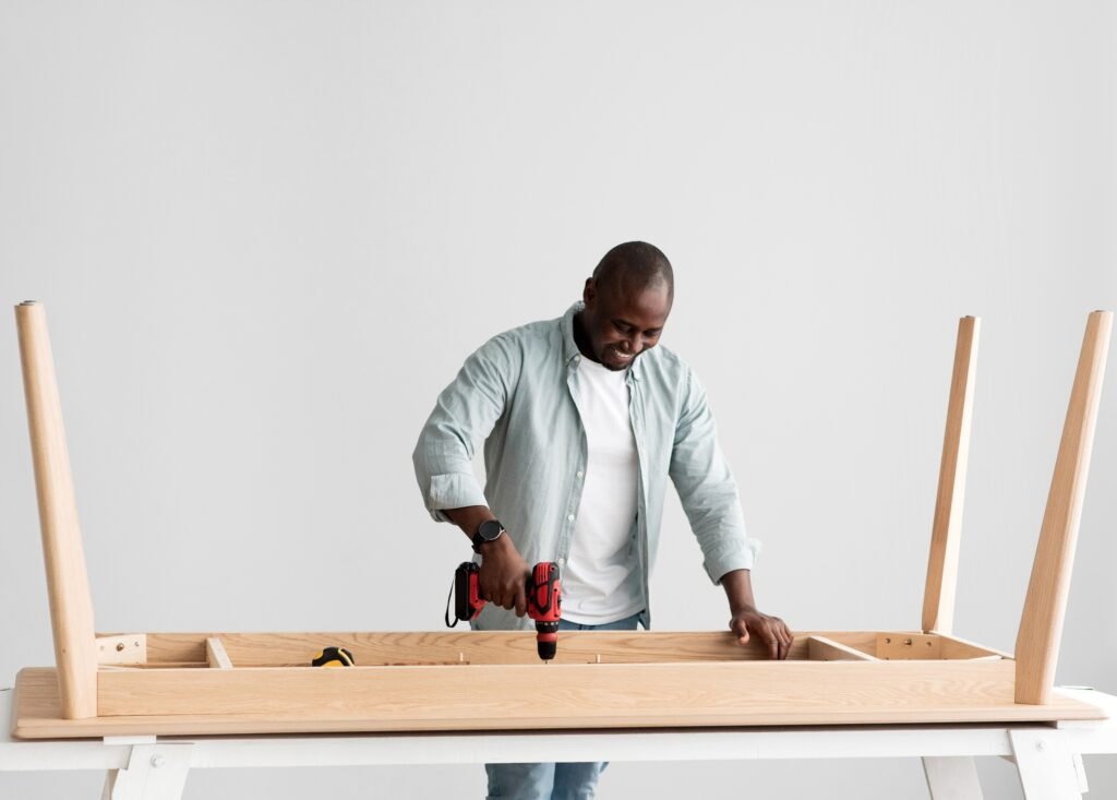 Carpentry service. Black handyman drilling wooden planks of table over light wall, free space