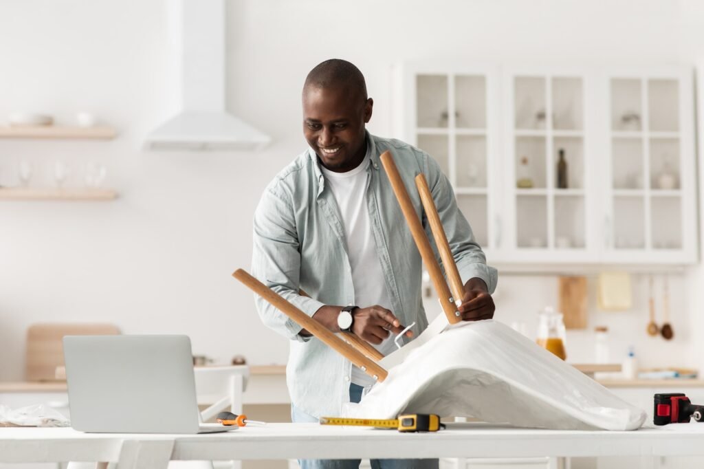 Furniture assembly. Happy african american man screwing detail to chair, looking at laptop at manual